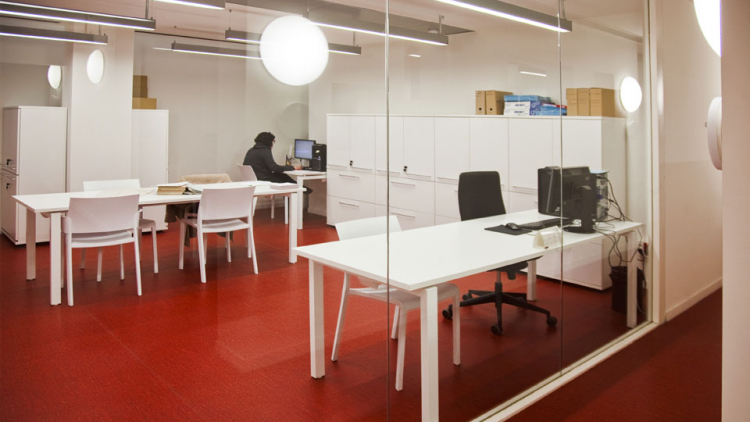 Imagen de la sala de consulta del AMDE, totalmente blanca, con mesas, armarios y una persona sentada en una de las mesas ante un ordenador