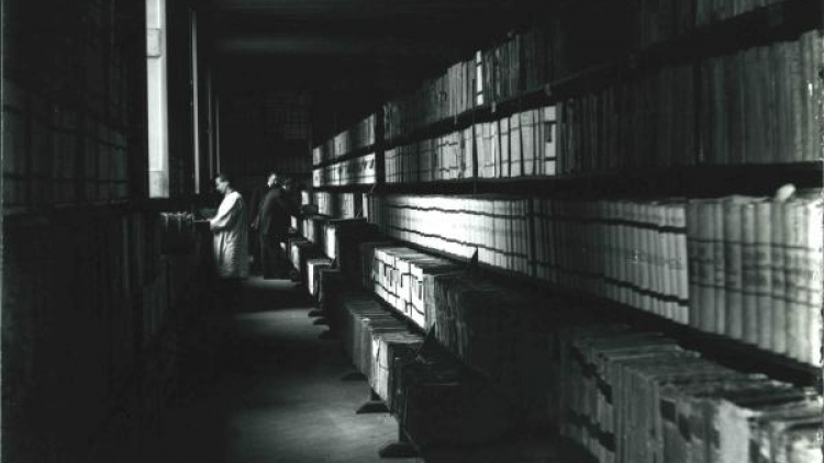 Fotografia en blanc i negre de l’interior del’arxiu als anys trenta, s’hi veuen dues persones, una amb una bata blanca, i tot de prestatges amb llibres