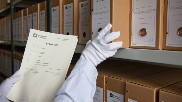Una mano con un guante blanco sujeta un documento mientras la otra, también enguantada, estira una caja de un estante