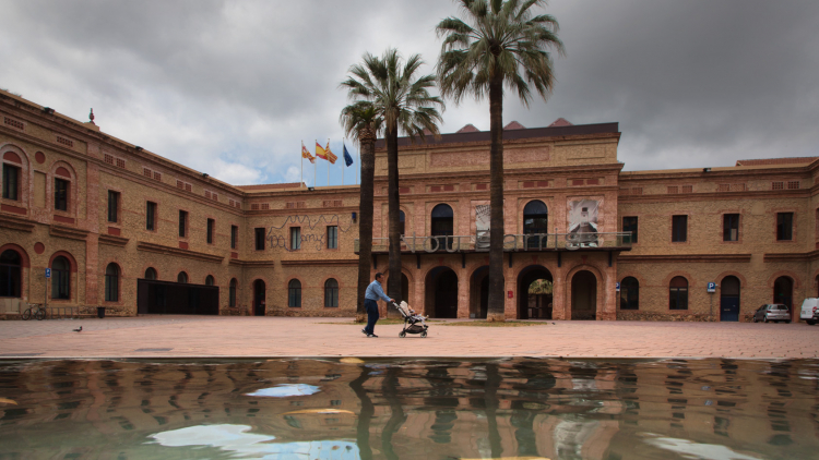 Image of a two-storey brick building, with a central body comprising a balcony and an entrance porch, which leads to a square with palm trees and a pond by which a person is walking.