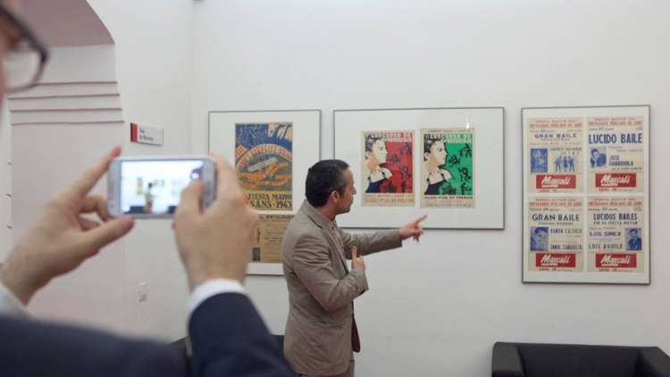 A primer pla es mostren unes mans que subjecten una càmera fotogràfica, la qual està prenent una imatge d'una persona que està explicant una sèrie d'obres als visitants d'una exposició.