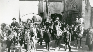 Four well-dressed men wearing hats on horses, which are held by other men. One of the men on horseback is holding a flag. All of them are in front of a large group of people and, in the background, there are some arcades and, on the right, the doorway of a church.