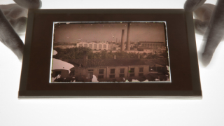 Dues mans sostenen una fotografia antiga d'una fàbrica del Districte de Sant Martí sobre una superfície lumínica.