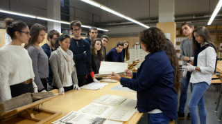 Un grup de persones escolten les explicacions d’una altra que té un document a les mans, al voltant d’una taula que mostra diversos documents