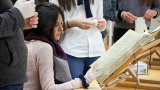 Una chica sentada mira un volumen manuscrito con otras personas a su alrededor