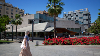Vista de l’edifici Jaume Fuster, que allotja l’AMDG i la biblioteca del districte de Gràcia, amb uns parterres de flors i palmeres al davant i persones caminant per la plaça