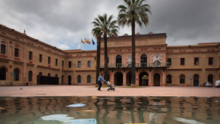 Image of a two-storey brick building, with a central body comprising a balcony and an entrance porch, which leads to a square with palm trees and a pond by which a person is walking.