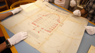 White gloved hands placed on top of, or on the verge of touching a plan showing the basilica ground plan of the Sagrada Família