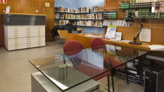 Vista de una sala de consulta con mesas, una de las cuales ocupada por una persona que consulta documentación, estanterías llenas de libros y un armario llano al fondo