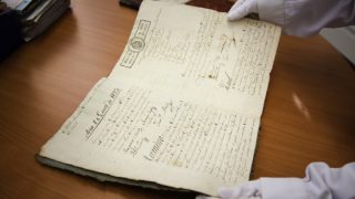 Unas manos con guantes blancos sostienen un legajo manuscrito abierto por la mitad 