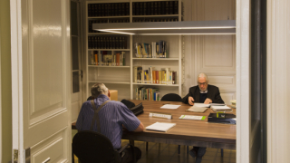 Dues persones estan assegudes en una taula consultant documentació, en una sala amb prestatgeries plenes de llibres