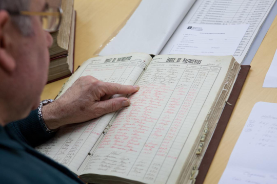 Un home d'edat avançada i amb ulleres consulta un llibre antic de naixements de Barcelona. L'home ressegueix amb el dit de la mà esquerra els registres que consten al llibre. 