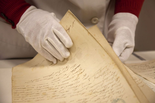 Unes mans amb guants subjecten documents antics manuscrits en un arxiu.