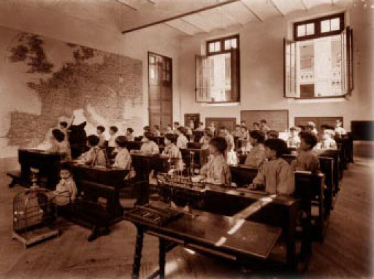 Tota una sèrie de nens es troben asseguts als seus escriptoris mentre observen, a mà dreta, un mapa d'Europa. Al fons hi ha dues finestres obertes.