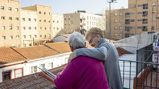 dos mujeres mayores se abrazan y se dan apoyo en una terraza con vistas a la ciudad de Barcelona